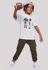 White cotton t-shirt with astronaut graphic, olive cargo pants, and white sneakers. Child holds a brown football, wearing a gray beanie.