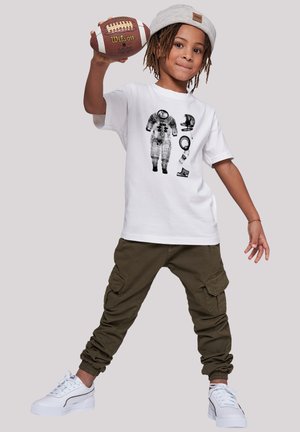 White cotton t-shirt with astronaut graphic, olive cargo pants, and white sneakers. Child holds a brown football, wearing a gray beanie.