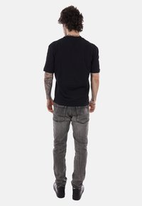 Hombre con cabello rizado, brazo derecho tatuado, vestido con camiseta negra, jeans grises y zapatillas negras, de pie de espaldas sobre fondo blanco.