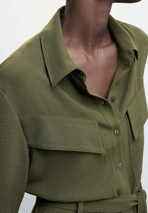 Chemise vert olive avec une finition texturée, dotée d'un col, de boutons sur le devant et de deux poches poitrine. Détail accentué à la taille ceinturée.