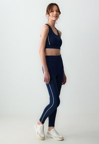 Top sportivo blu navy e leggins con accenti bianchi ai lati, realizzati in tessuto elasticizzato. Scarpe da ginnastica bianche con accenti beige chiaro.