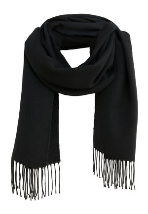Femme debout contre un mur beige portant un cardigan argyle gris et vert, une jupe noire, une écharpe noire et des bottes noires mi-mollet.