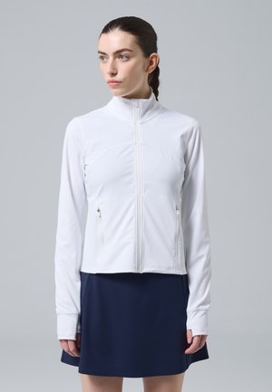 Donna con capelli scuri raccolti in una coda di cavallo, che indossa una giacca bianca con cerniera e una gonna blu navy, guarda verso sinistra su uno sfondo grigio uniforme.