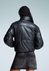 Oseba z dolgimi pletenicami, oblečena v črno usnjeno puhovko in temno jeans mini krilo, stoji obrnjena stran z rokama na bokih.