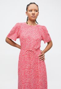 Κόκκινο φόρεμα με λευκό πουά σχέδιο, κοντά φουσκωτά μανίκια, στρογγυλή λαιμόκοψη και ζώνη με κορδόνι στη μέση. Ομαλή υφή υφάσματος.