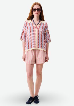 Femme portant un polo en tricot à rayures oversized, un short à carreaux rose, des baskets noires, un collier de perles bleu, et de grandes lunettes de soleil foncées.