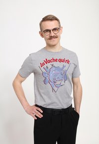 Šedé bavlněné tričko s grafikou "La Vache qui rit" v červených a modrých linkách, s krátkými rukávy, kulatým výstřihem a volným střihem, spárované s černými kalhotami.