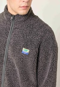 Vyras dėvi tamsiai pilką tekstūruotą fliso striukę su priekyje esančiu užtrauktuku ir mažu spalvotu lipduku kairiajame krūtinės skyriuje, kuriame parašyta „La Maison AMV“.