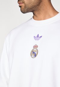 Λευκό φούτερ με κεντημένο μωβ λογότυπο Adidas trefoil και έμβλημα της Ρεάλ Μαδρίτης με στέμμα, που παρουσιάζει ομαλή υφή και σχεδιασμό με στρογγυλή λαιμόκοψη.