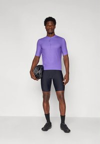 Sykleantrekk med en lys lilla sykkeltrøye med korte ermer, svarte shorts, svart hjelm i hånden og svarte sykkelsko.