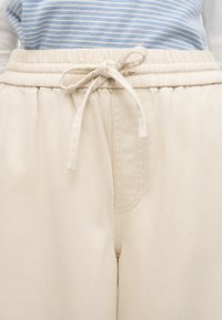 Κοντινό πλάνο σε μπεζ παντελόνι με ελαστική μέση και κορδόνι δεμένο σε φιόγκο, συνδυασμένο με μπλε και λευκή ριγωτή μπλούζα.