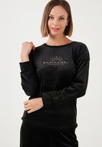 Zwart long sleeve top van zachte fluweel met een ronde halslijn. Kenmerken zijn de gouden tekst "sunrise candle co" en een subtiele afgeronde zoom.