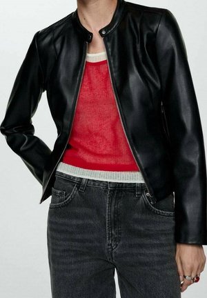 Person trägt eine schwarze Lederjacke über einem rot-weißen Pullover, kombiniert mit dunkelgrauen, hoch taillierten Jeans und Ringen an der rechten Hand.