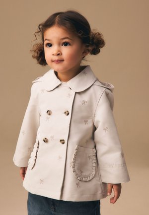 Junges Kind mit lockigem Haar, das einen beigen zweireihigen Mantel mit Schleifenmustern und Rüschen an den Taschen trägt, steht vor einem schlichten Hintergrund.