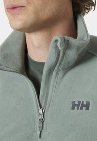 Fleeceová bunda ve světle zelené barvě s límcem na zip, se černým logem na hrudi a hladkým povrchem. Ucho zipu je stříbrné.