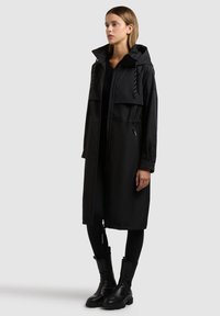 Ženska v dolgem črnem plašču s kapuco, zoženim v pasu, črnih legicah in črnih gležnjarjih, stoji pred enobarvnim sivim ozadjem.