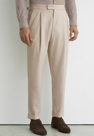 Mężczyzna ubrany w beżowe plisowane spodnie, brązową koszulę w paski, brązowe skarpetki i ciemnobrązowe zamszowe mokasyny, stojący przy białej ścianie.