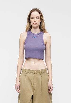 Fiatal nő hosszú szőke hajjal, ujjatlan lila crop toppal és bézs bő szárú nadrággal, egyszínű háttér előtt állva.