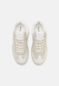 Hvite og beige sneakers laget av semsket skinn og lær, med rund tå, snøring, og en logo på tungen. Teksturierte detaljer overalt.