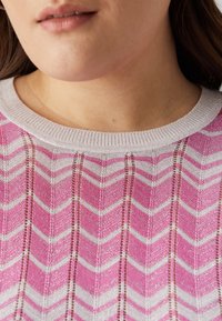Růžová a bílá pletenina s chevron vzorem, s texturovaným povrchem a jemným leskem; má žebrovaný kulatý výstřih a detaily s otevřeným tkaním.