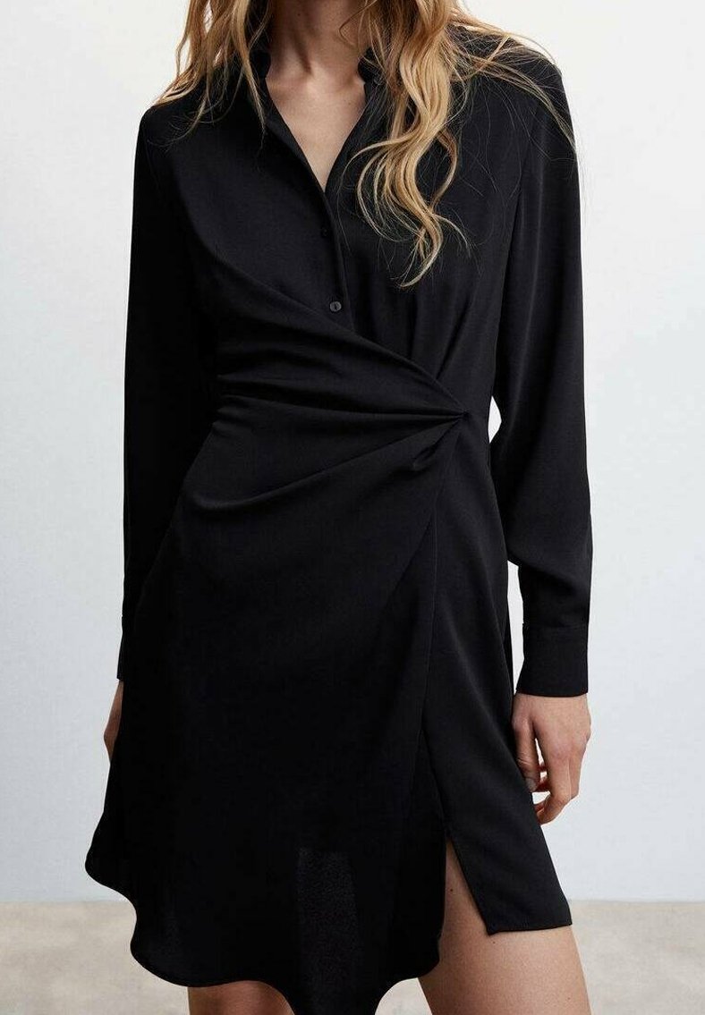 Femme portant une robe noire à manches longues avec un détail froncé à la taille et une fente latérale, debout devant un fond clair uni.