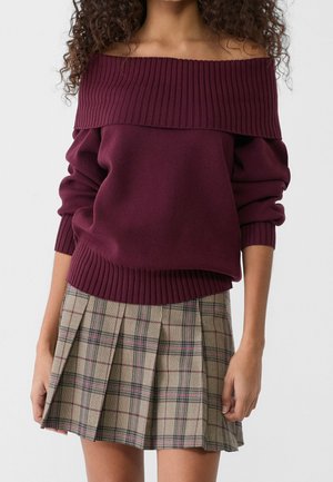 Woman wearing a burgundy off-shoulder ribbed sweater and a beige plaid pleated skirt against a plain background.