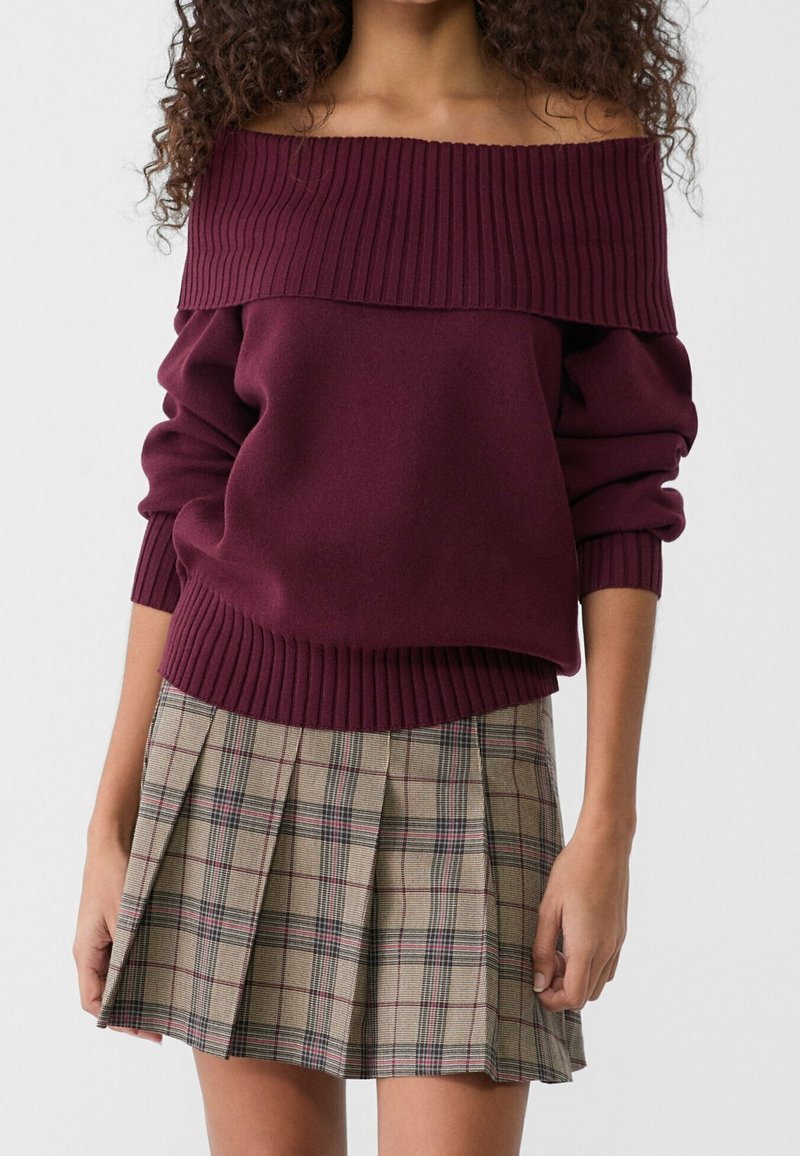 Femme portant un pull à épaules dénudées côtelé bordeaux et une jupe plissée à carreaux beige sur un fond uni.