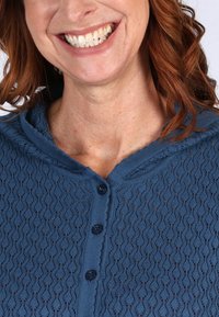 Blauer gestrickter Cardigan mit einem strukturierten Wellenmuster, sechs dunklen Knöpfen auf der Vorderseite und einem runden Kragen. Weiches Material, mittlere Gewicht.