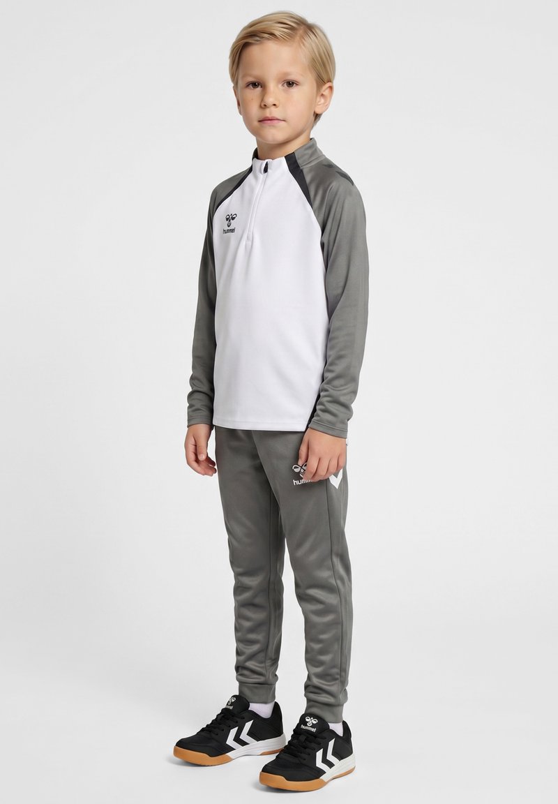Blond jongetje staat, draagt een witte en grijze sportjas, grijze broek en zwarte sneakers met witte chevron-logo's.