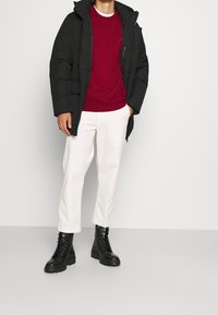 Cappotto imbottito nero, maglione rosso a collo rotondo, pantaloni crema e stivali neri stringati. Il cappotto presenta un cappuccio e tasche frontali.