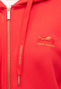 Raudona zip-up gobtuvu striukė, pagaminta iš minkštos medžiagos, su auksiniu logotipo siuvinėjimu ir suderintomis raištelių detalėmis. Auksinė užtrauktuko fiksavimo detalė.