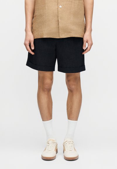 Homme portant un short noir retroussé, des chaussettes blanches à côtes, des baskets beige et blanches, et une chemise beige boutonnée, vu de la taille jusqu'aux pieds.