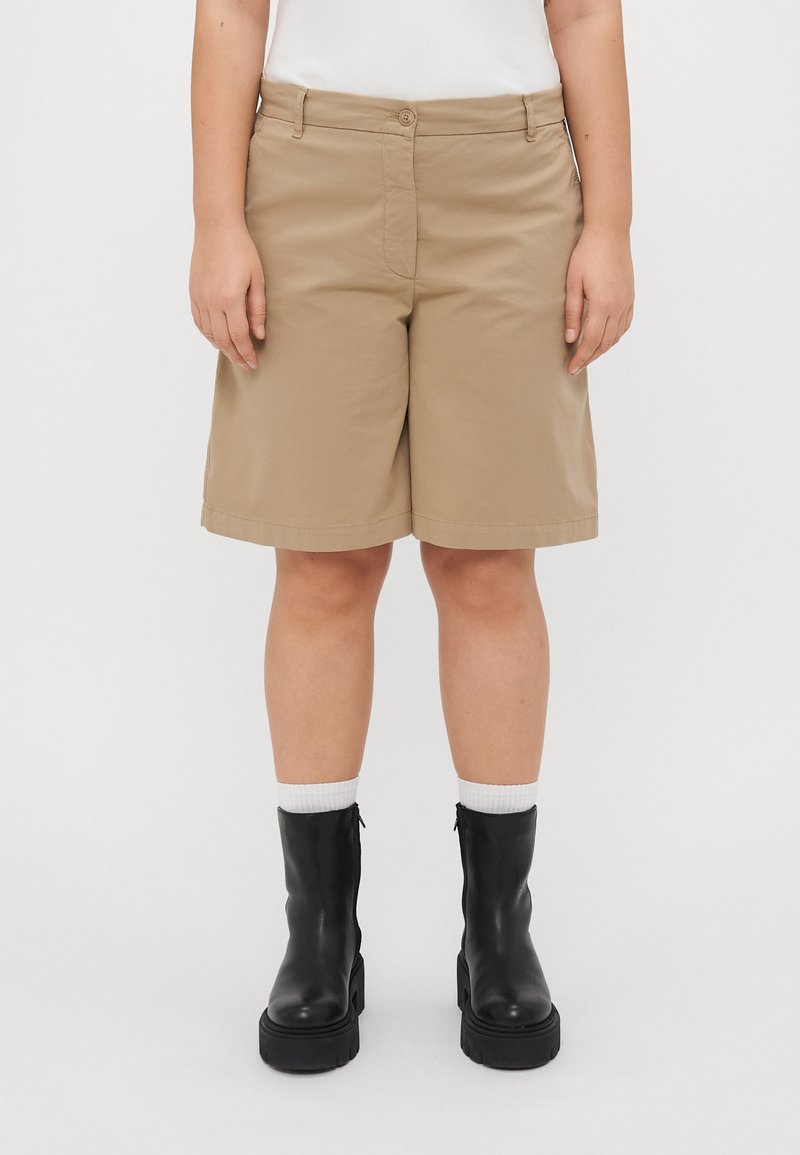 Beige Shorts mit mittellangem Design, aus glattem Material, mit Knopfverschluss und Gürtelschlaufen, kombiniert mit schwarzen Stiefeletten.