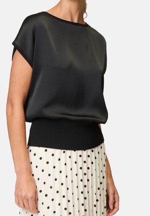 Femme portant un haut noir sans manches avec une large bordure côtelée et une jupe plissée blanche à pois noirs, sur fond blanc.