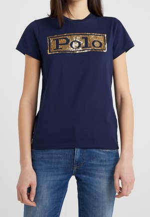 Person iført en mørkeblå t-shirt med guld paliettekst "Polo" på brystet og blå jeans mod en ensfarvet baggrund.