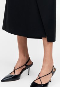 Γυναίκα φοράει μαύρες μυτερές γόβες slingback με λεπτούς σταυρωτούς ιμάντες και μαύρη φούστα μεσαίου μήκους.