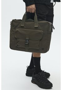 Sac en nylon vert olive avec deux poignées supérieures, poche à rabat avant sécurisée par une boucle, fermeture éclair et détail de marque sur le côté.