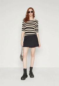 Woman with long red hair wearing round sunglasses, black-and-white striped top, black mini skirt, black boots, and holding a small gray handbag.