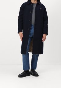 Cappotto di lana blu navy con una texture liscia, caratterizzato da un colletto, chiusura con bottoni e rivestimento a contrasto, abbinato a jeans blu e scarpe nere.