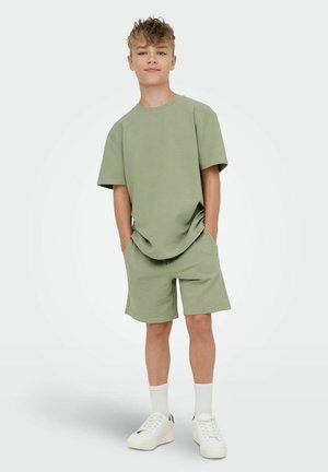 Dreng står med hænderne i lommerne iført lysegrøn tekstureret kortærmet skjorte og matchende shorts, hvide sokker og hvide sneakers.