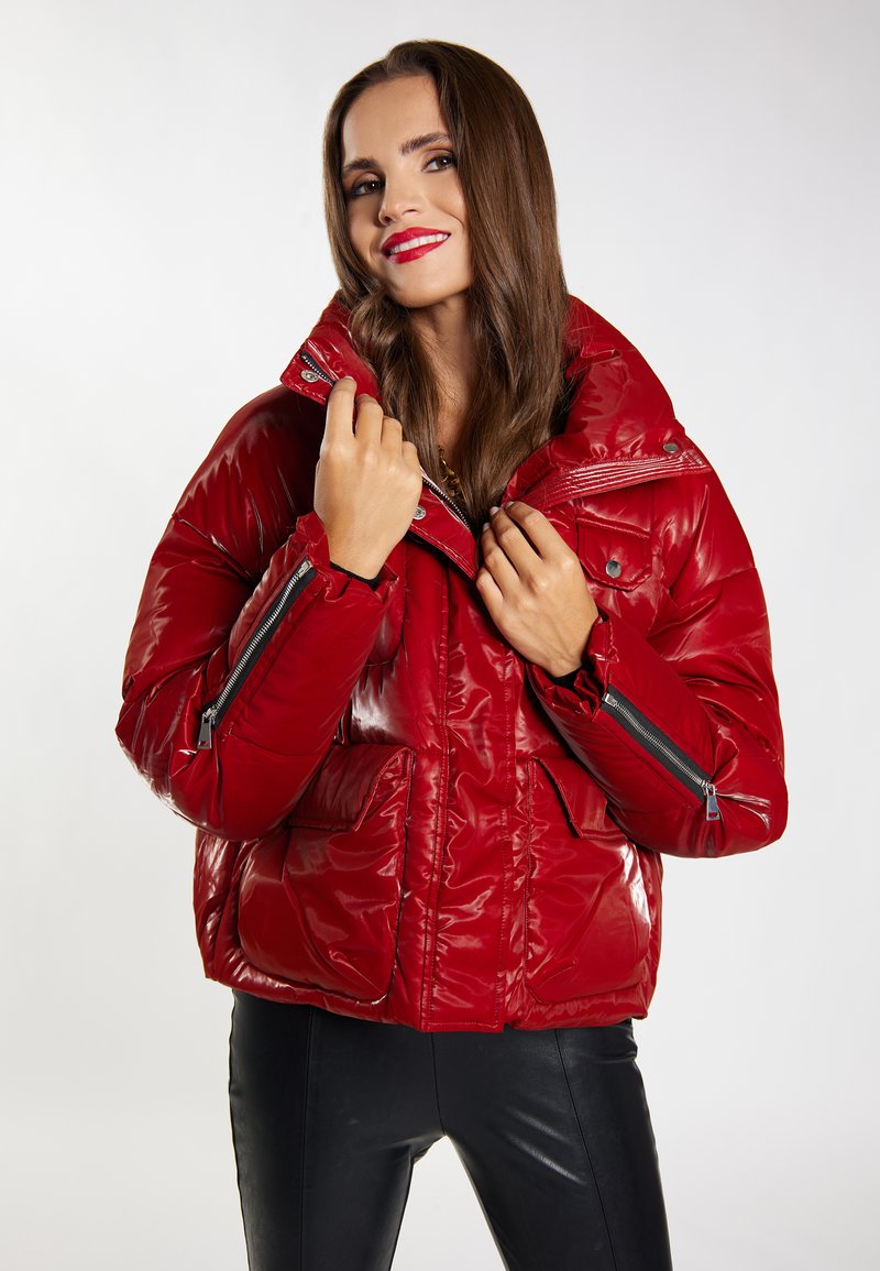 Giacca imbottita rossa con finitura lucida, colletto alto e tasche con zip; presenta una vestibilità rilassata e dettagli di cucitura in evidenza.