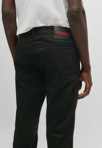 Pantalon en denim noir avec une coupe droite, doté de poches arrière et d'une patch en cuir noir de marque avec le lettrage rouge "HUGO" sur la ceinture.
