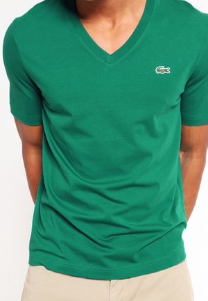 T-shirt en coton vert avec col en V et manches courtes, arborant un petit logo brodé sur le côté gauche de la poitrine et une texture lisse.