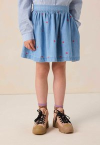 Jupe en denim avec taille froncée, couleur bleu clair et motifs de cœurs rouges. Associée à des baskets beiges épaisses et des chaussettes à motifs.