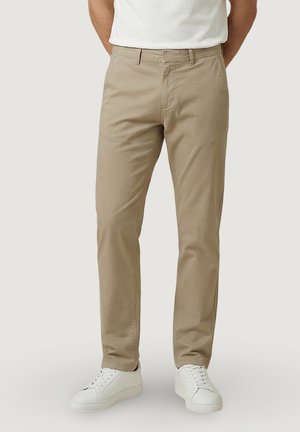 Homme portant un pantalon beige à jambes droites, des baskets blanches et une chemise blanche à manches courtes rentrée, debout les mains derrière le dos.