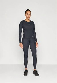 Egy nő áll, fekete hosszú ujjú sportfelsőben és leggingsben, fekete futócipővel egy sima világos háttér előtt.