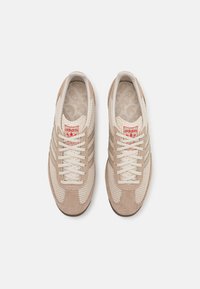 Zapatillas deportivas beige con parte superior de malla, acentos de ante, cordones beige y tres franjas icónicas en crema a los lados. Logo rojo en la lengüeta.
