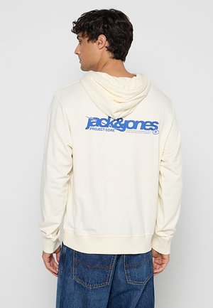 Férfi, aki krémszínű kapucnis pulóvert visel kék „jack & jones Project Core“ felirattal a hátán, kék farmernadrággal párosítva, oldalra néz.