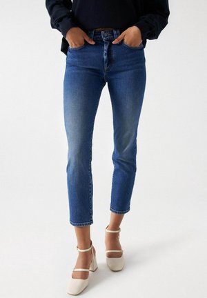 Person iført cropped blå denim jeans og hvide Mary Jane sko med blokhæl, hænder i lommer, stående mod hvid baggrund.