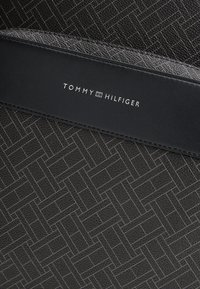 Μαύρο υλικό με υφή και μοτίβο ψαροκόκκαλο· διαθέτει λείο μαύρο δερμάτινο λουρί με ανάγλυφη την επιγραφή "TOMMY HILFIGER" σε λευκό χρώμα.